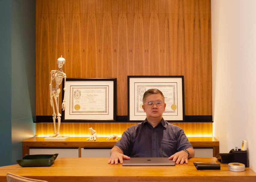 Tomas Makiyama está sentado na mesa da sua sala, com as mãos sobre a mesa onde se encontra um notebook fechado, ao lado alguns itens de escritório e ao fundo dois quadros com diplomas e um esqueleto decoram a sala.